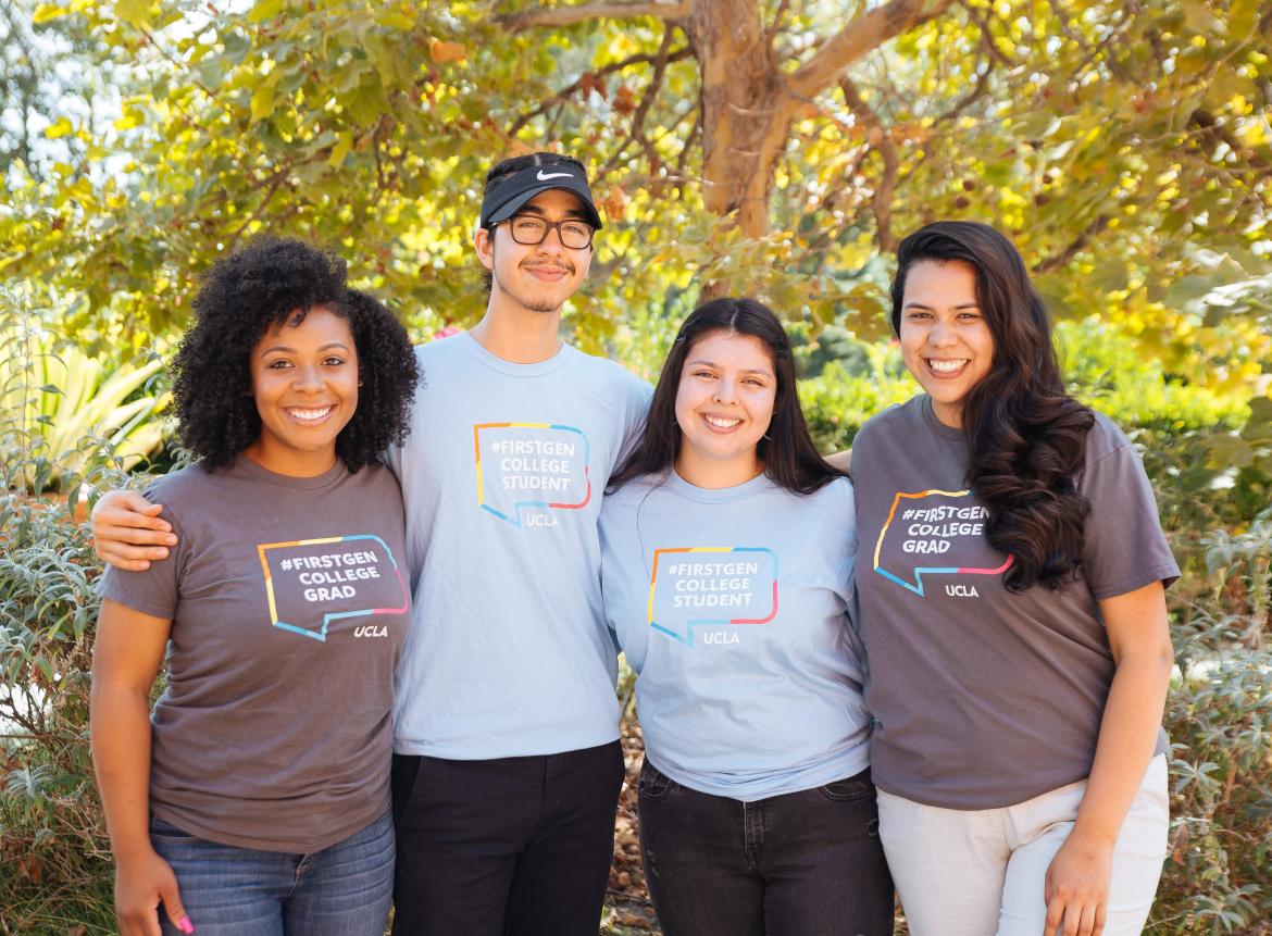 Picture of four students standing in front of a tree, smiling and proudly wearing a #First-Gen College Grad and First Gen College Student Shirt.